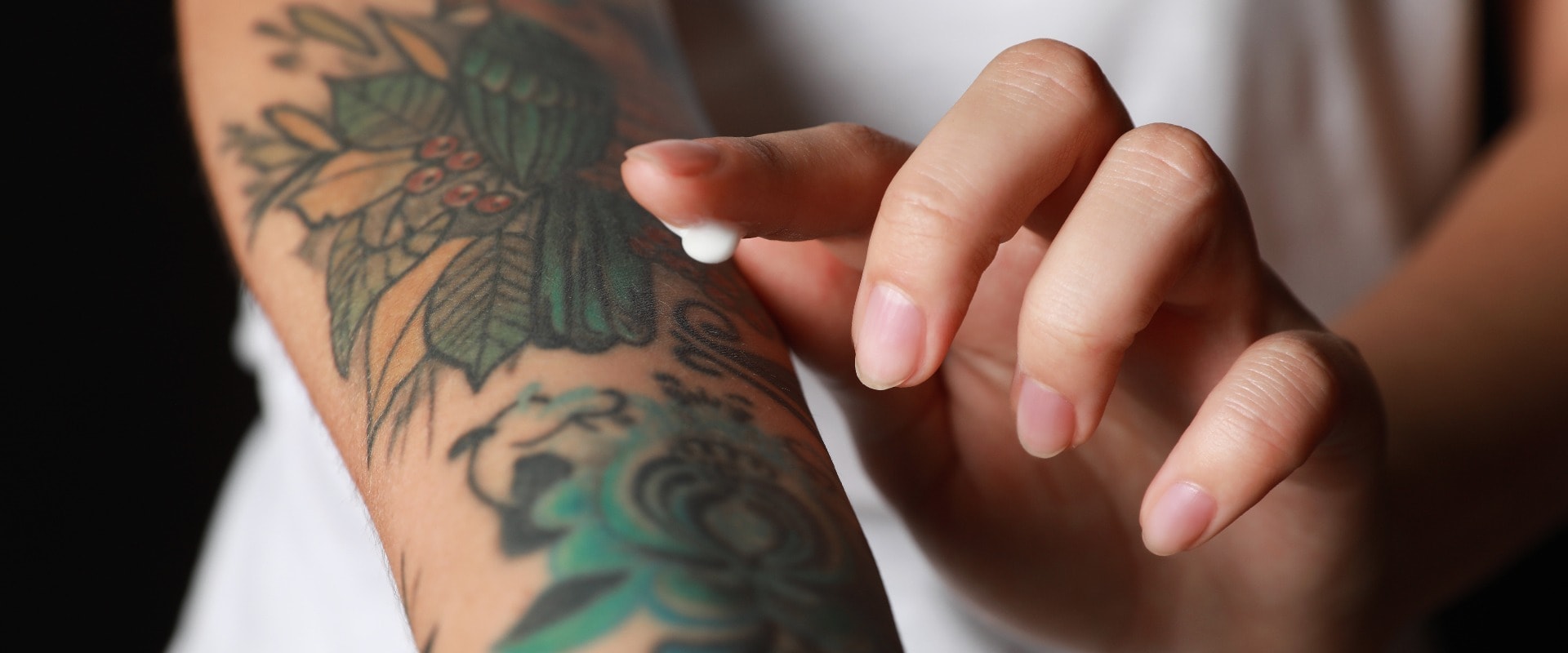 Woman applying cream on her arm with tattoos against black background, closeup